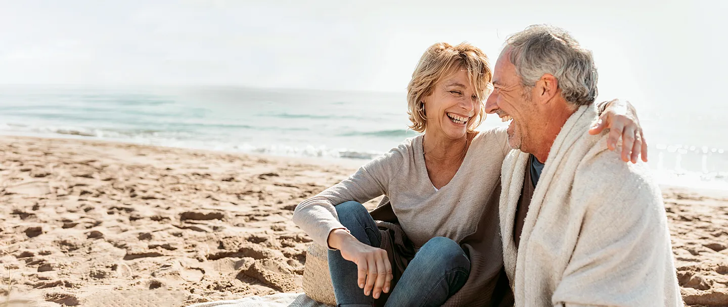Paar umarmt einander während es am Strand auf einer Picknickdecke sitzt.